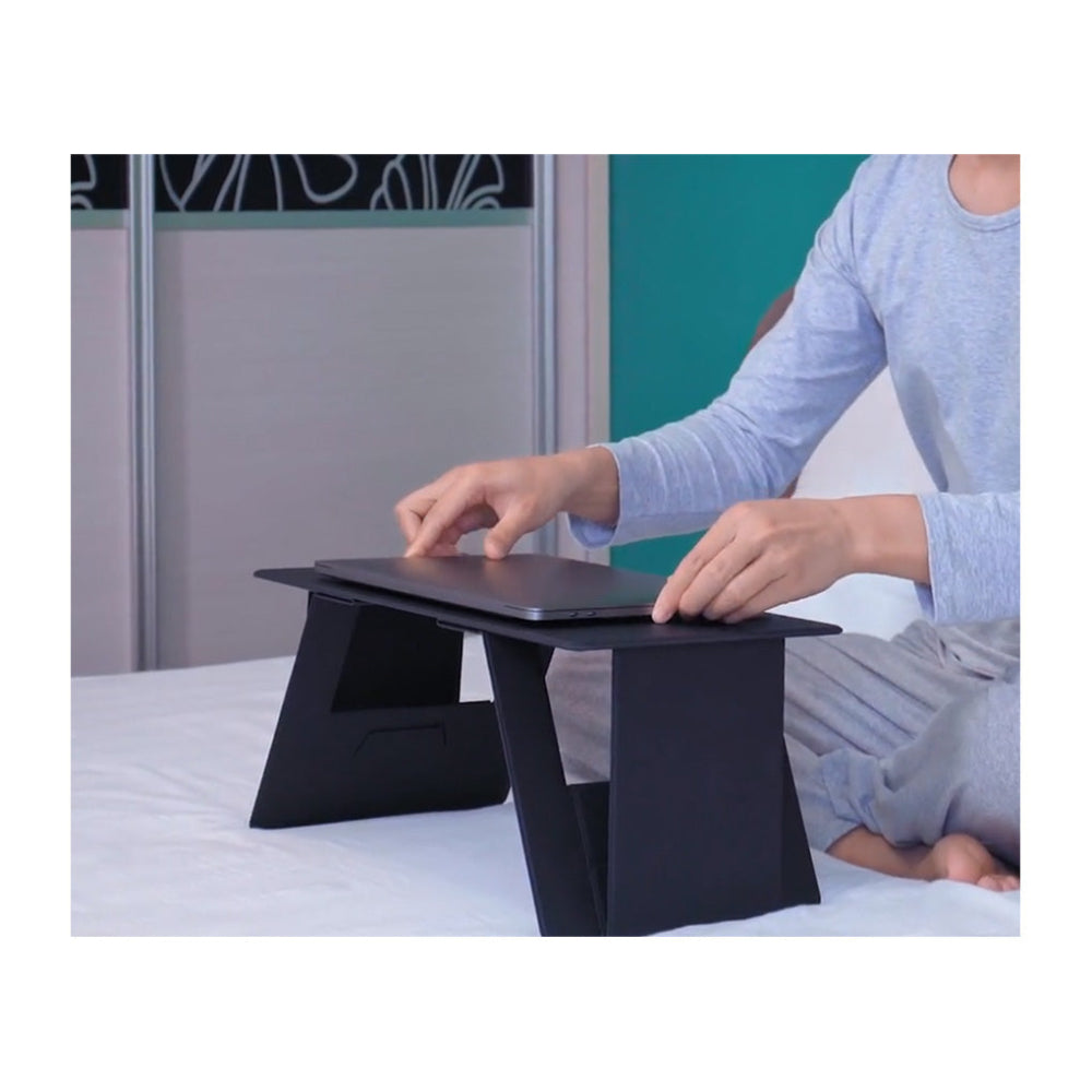 Person using a black adjustable stand to work on a laptop on a bed.